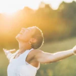 A woman with short hair smiling with their arms outstretched in a sunlit field, feeling balanced and in harmony with the season's energy.