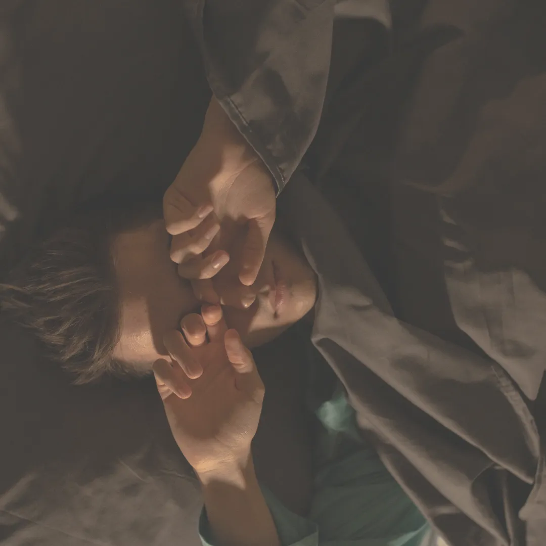 Man lying in bed under dark sheets, rubbing his eyes before sleep, illustrating nighttime rest and recovery