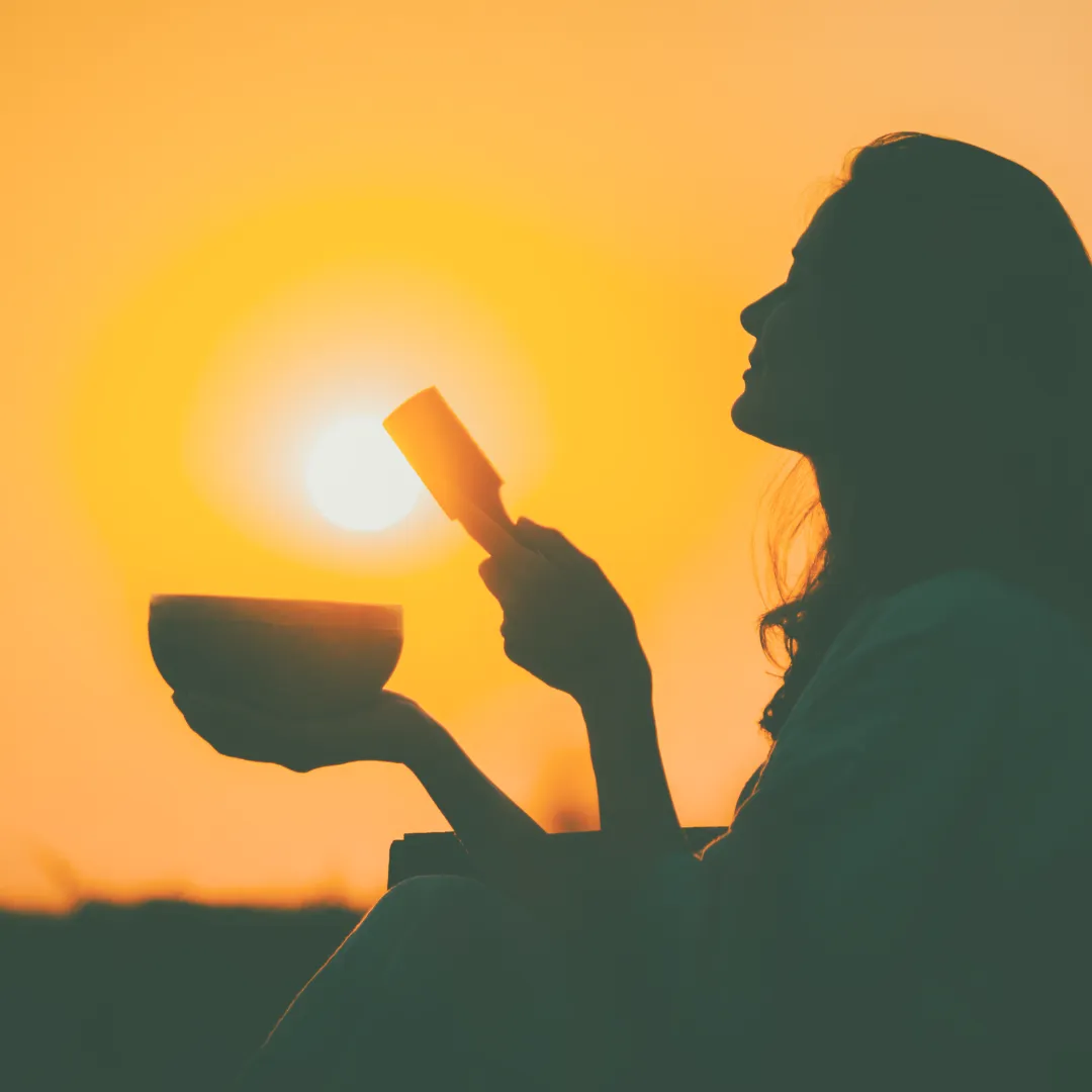 Silhouette of a woman practicing sound healing meditation with a singing bowl as the sun sets behind her.