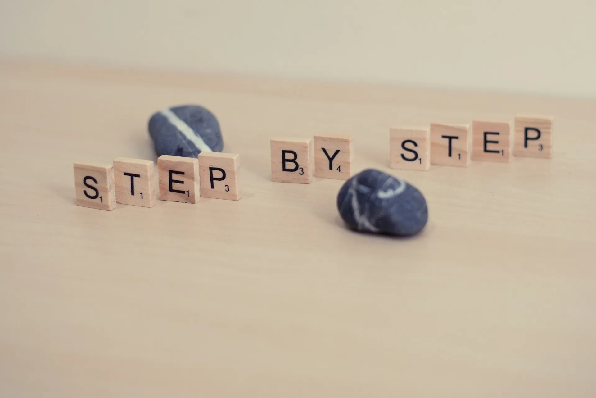 Wooden letter tiles spelling step by step beside two smooth grey stones on a pale surface