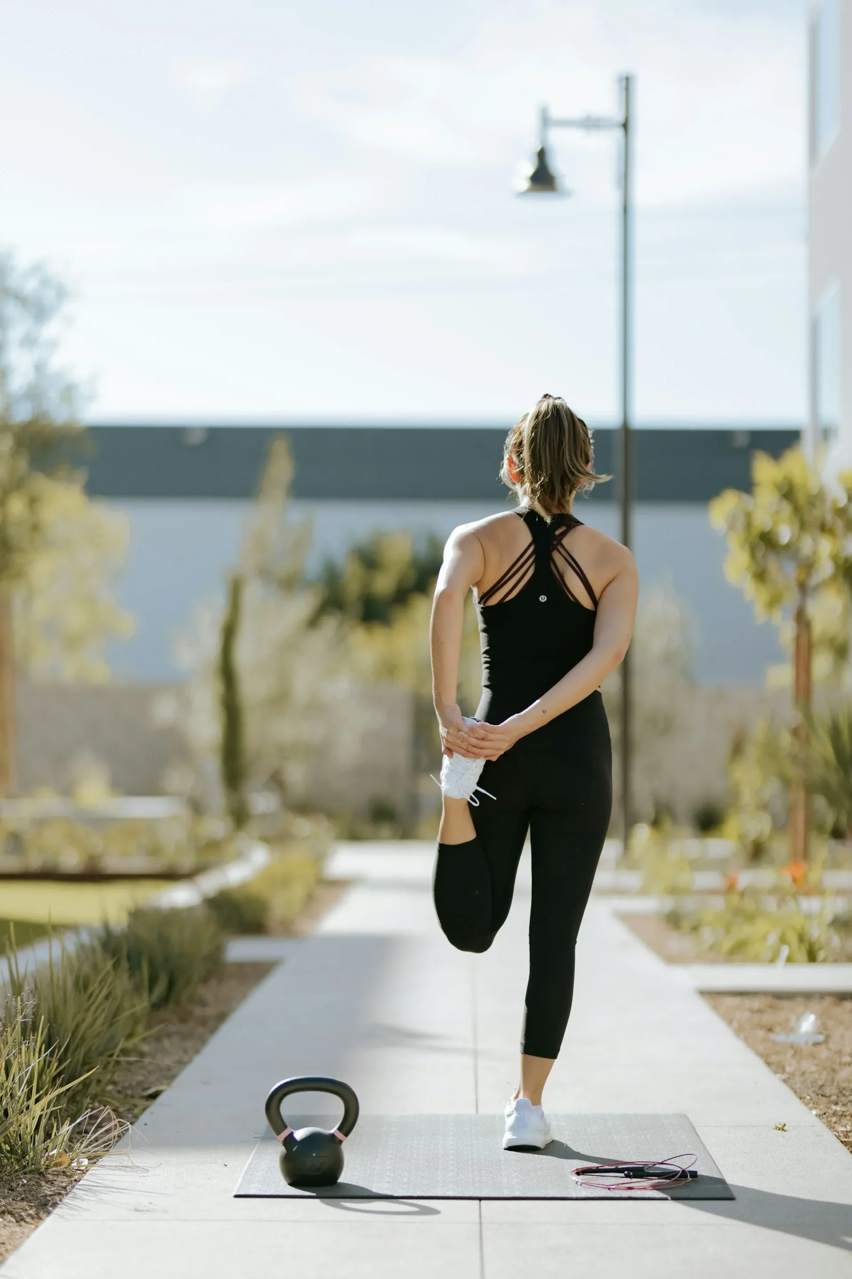 Woman stretching on an exercise mat outdoors beside a kettlebell and skipping rope