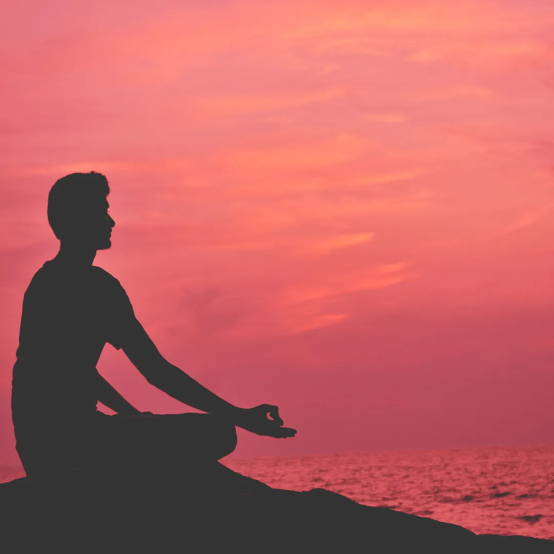 Man meditating in lotus position at sunset by the ocean, symbolizing mindfulness and inner peace.