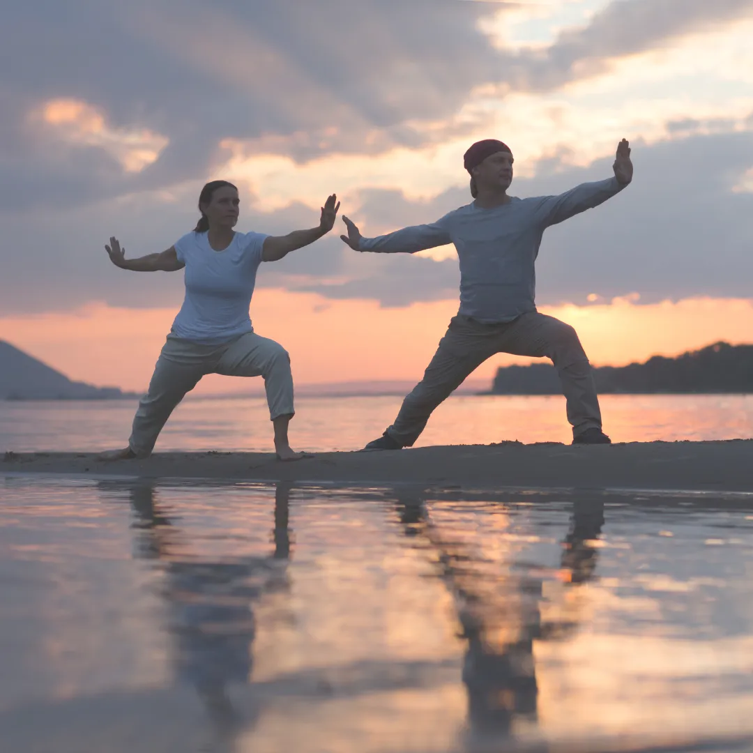 A couple practicing Tai Chi on the beach at sunset, engaging in mindful movement for detoxification and energy balance.