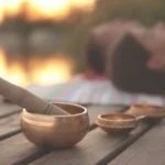 A brass Tibetan singing bowl rests on a wooden deck during a sound bath, with a person relaxing on a mat in the background near a warm light.