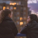 Two friends leaning on a railing talking at dusk with city lights behind them