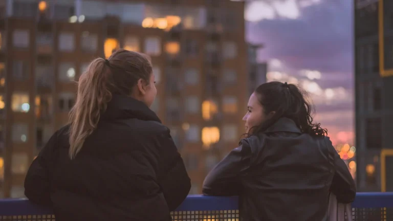 Two friends leaning on a railing talking at dusk with city lights behind them