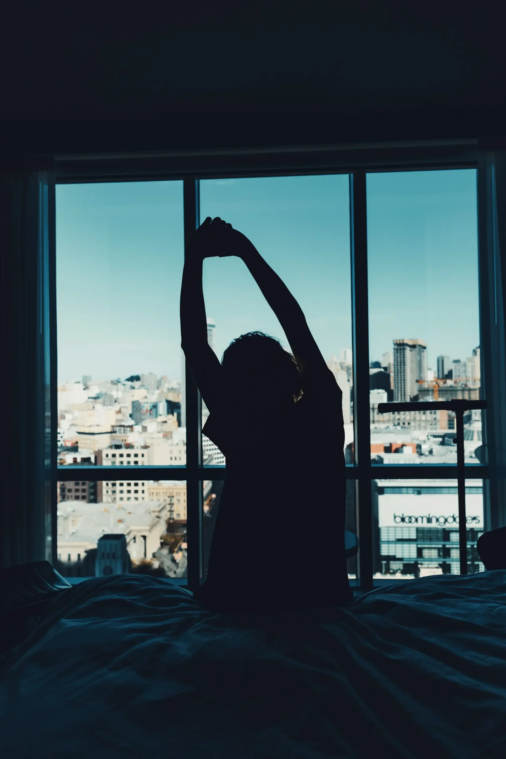 Silhouette of a person stretching while sitting up in bed in front of a city window at dawn