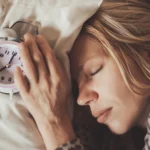 A woman sleeping peacefully next to a retro alarm clock on a pillow, illustrating the importance of aligning sleep schedules with your chronotype.
