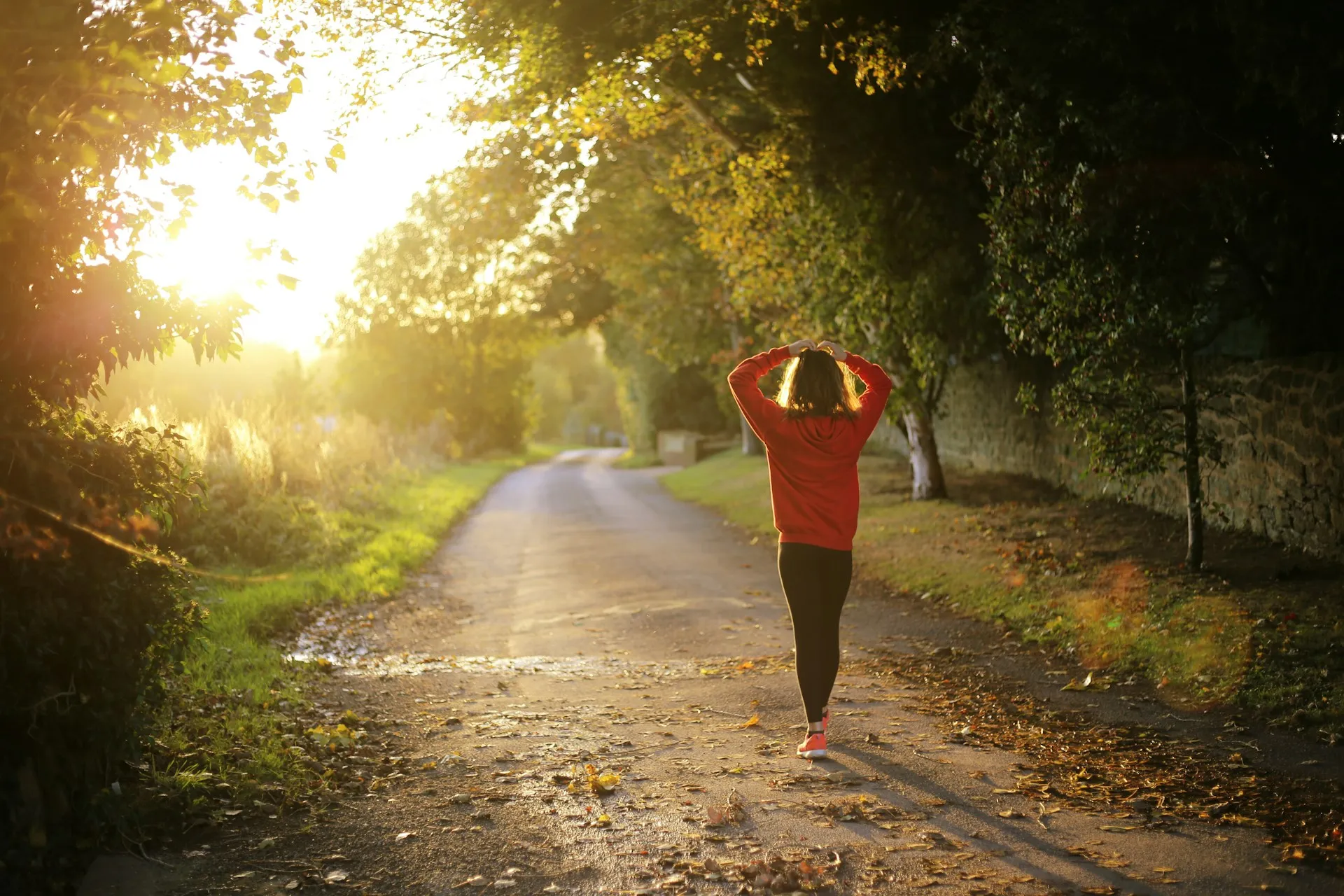 Woman in a red sweatshirt walking along a tree-lined road in warm evening sunlight