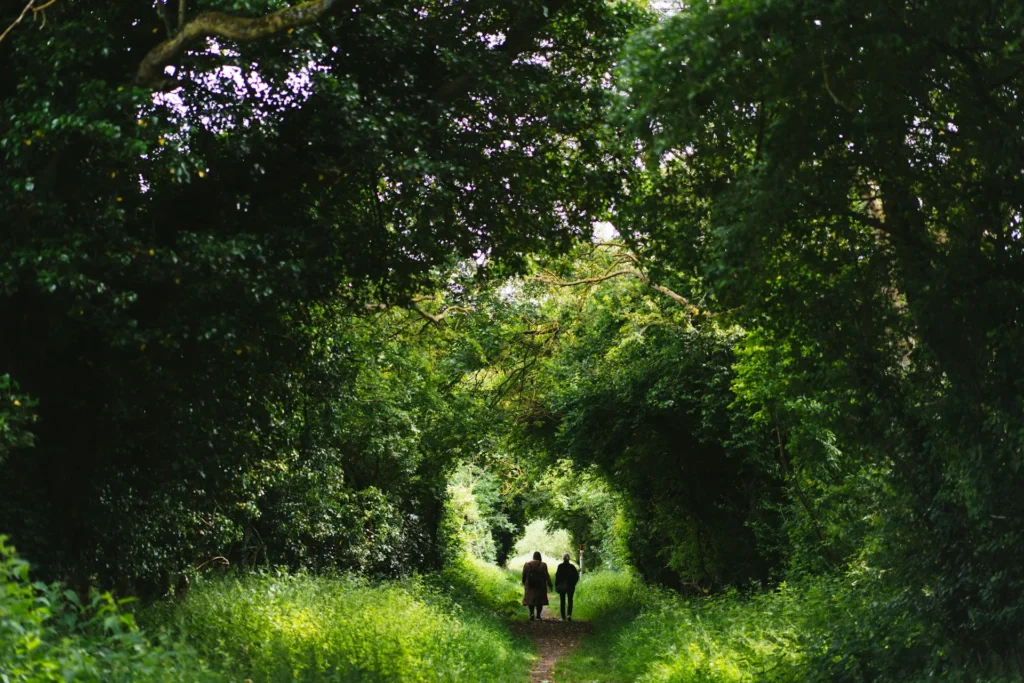 Two people walking together along a narrow path through a lush green tree tunnel