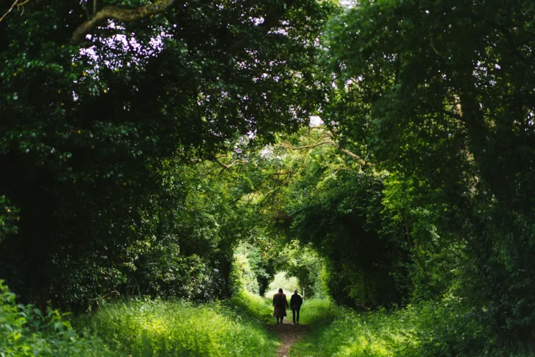 Two people walking together along a narrow path through a lush green tree tunnel