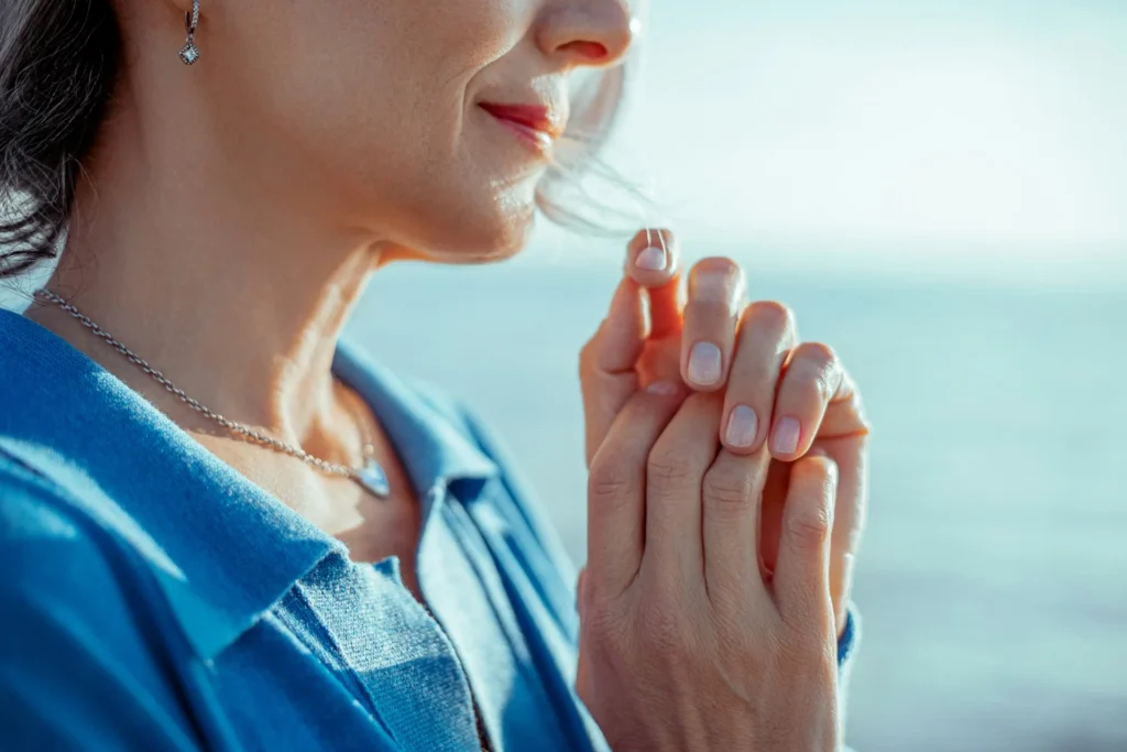 Woman standing outdoors by the water with her hands clasped near her chest, eyes lowered
