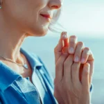 Woman standing outdoors by the water with her hands clasped near her chest, eyes lowered