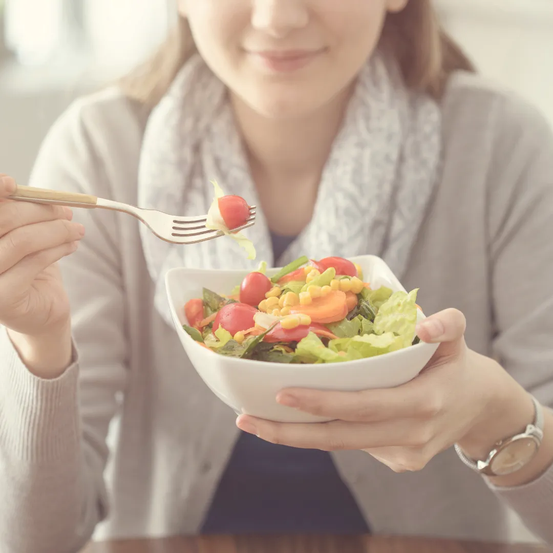 A young woman smiling as she eats a fresh vegetable salad from a white bowl, containing lettuce, corn, and cherry tomatoes.