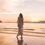 Woman in a flowing white dress walking barefoot on a wet sandy beach at sunset, practicing mindful walking for stress relief.
