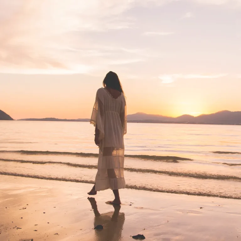 Woman in a flowing white dress walking barefoot on a wet sandy beach at sunset, practicing mindful walking for stress relief.