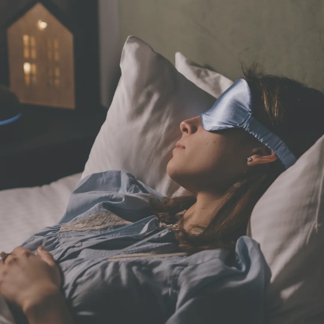 Woman wearing a silky blue sleep mask while lying in bed, promoting deep sleep hygiene and recovery